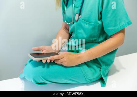 Immagine irriconoscibile ritagliata di un'infermiera in scrub verdi seduta su una scrivania, utilizzando uno smartphone, rilassandosi durante una pausa in un set di studio medico Foto Stock