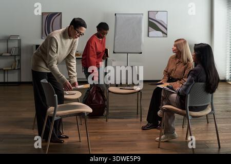 Gruppo di giovani adulti e donne multietniche di mezza età che si preparano alla psicoterapia di gruppo, organizzano sedie e tengono quaderni Foto Stock