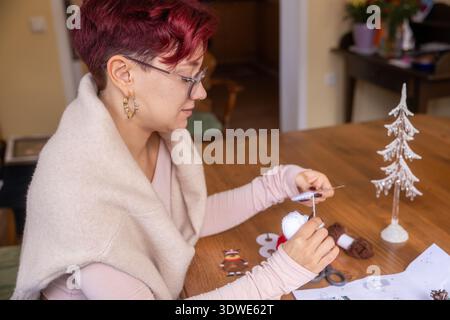 Una giovane donna con i capelli rossi corti si gode una giornata accogliente a casa, impegnandosi in una rilassante attività artigianale su un tavolo di legno, circondato da filati e festivi Foto Stock
