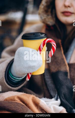 Una donna con un caldo abbigliamento invernale regala una tazza di caffè giallo adornata con una canna da zucchero. Le piace la sua bevanda calda all'aperto, avvolto in un abbigliamento accogliente per Foto Stock