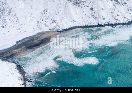 Vista aerea del litorale invernale delle Lofoten, con onde turchesi che si infrangono dolcemente contro la spiaggia innevata, che racchiude la regione in un ambiente sereno Foto Stock