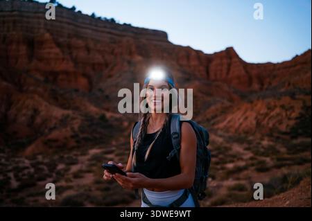 Una donna fa un'escursione attraverso il Red Canyon spagnolo, utilizzando il suo smartphone per esplorare il paesaggio mozzafiato. È dotata di un faro e di uno zaino, emp Foto Stock