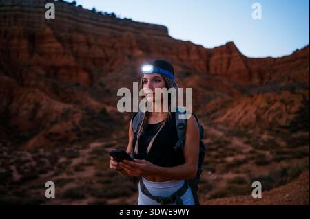 Una donna fa un'escursione attraverso il Red Canyon spagnolo, utilizzando il suo smartphone per esplorare il paesaggio mozzafiato. È dotata di un faro e di uno zaino, emp Foto Stock