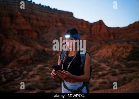 Una donna fa un'escursione attraverso il Red Canyon spagnolo, utilizzando il suo smartphone per esplorare il paesaggio mozzafiato. È dotata di un faro e di uno zaino, emp Foto Stock