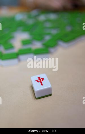 Primo piano di una piastrella mahjong con carattere rosso su un tavolo circondato da piastrelle verdi e bianche. Cattura un momento di gioco strategico in questo tradizionale Foto Stock