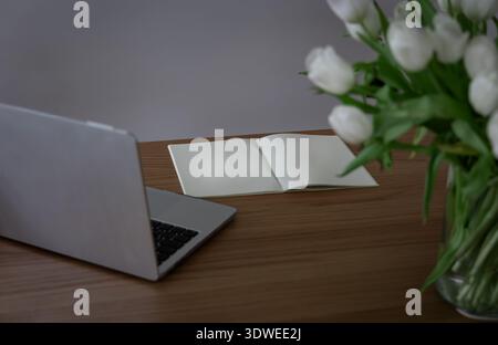 Spazio di lavoro minimo per l'ufficio domestico con laptop, notebook aperto e tulipani su una scrivania in legno, lavoro remoto e concetto di produttività. Foto Stock