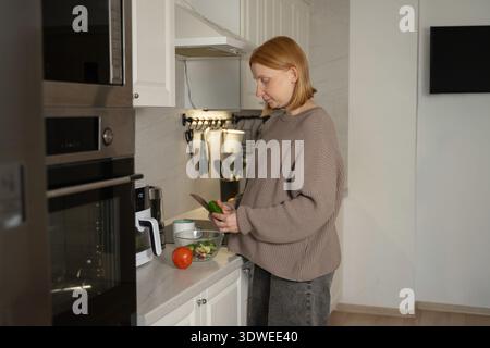 Una donna incinta si vede preparare un pasto sano nella sua cucina moderna, tritare verdure e garantire una dieta nutriente per lei e la sua ba in crescita Foto Stock