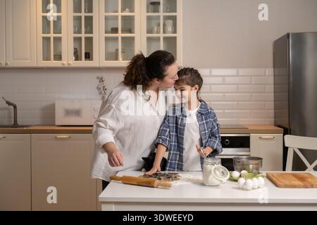 Una madre amorevole che bacia sua figlia sulla testa mentre si godono una divertente sessione di cucina nella loro accogliente cucina casalinga. La scena è piena di calore e Foto Stock