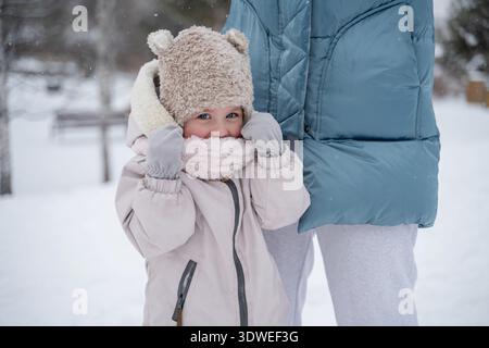 Un bambino vestito con un caldo cappotto, un cappello e una sciarpa si erge accanto a un genitore in un parco invernale innevato, con un'espressione giocosa e allegra. Foto Stock