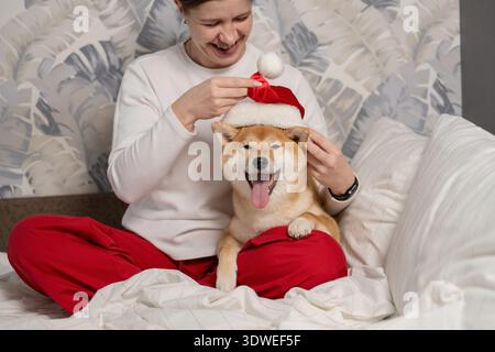 Una donna gioiosa siede comodamente su un letto, vestendo il suo felice Shiba Inu con un festoso cappello di Babbo Natale. La scena irradia calore, allegria natalizia e compagno di viaggio Foto Stock
