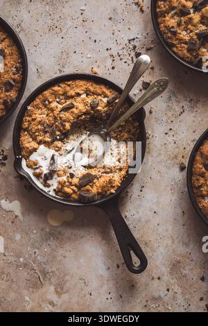 In una padella di ghisa viene servito un biscotto caldo e gustoso ripieno di pezzi di cioccolato, parzialmente mangiato con due cucchiai e un filo di panna e cappellino Foto Stock