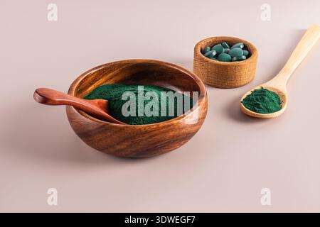 Spirulina in polvere in una ciotola di legno con un cucchiaio, accompagnata da compresse in una ciotola più piccola e più polvere su un cucchiaio. Toni caldi su un backgroun neutro Foto Stock