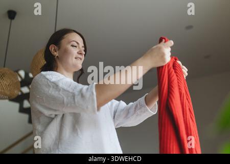 Una donna sorridente in un ambiente accogliente regge con cura un maglione rosso vibrante, esaminandone i dettagli. Gli interni moderni offrono un ambiente caldo e accogliente Foto Stock