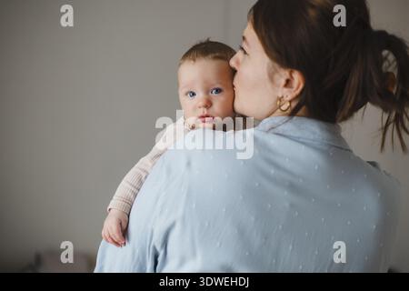 Un momento tenero mentre una madre bacia dolcemente il suo bambino appena nato mentre lo tiene vicino. Il bambino guarda curiosamente la fotocamera, avvolto in un'amata cornice Foto Stock