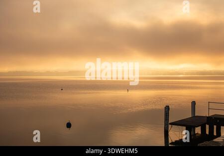 Una tranquilla scena al tramonto con una tonalità dorata che si estende su un lago calmo, che riflette le dolci nuvole sopra. La quiete è punteggiata da un piccolo dottore Foto Stock