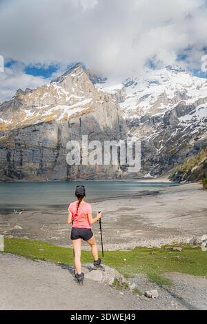 Un escursionista con un abbigliamento vivace si erge sullo splendido lago Oeschinen, annidato nelle Alpi svizzere. Le cime innevate e le foreste lussureggianti forniscono un pict Foto Stock
