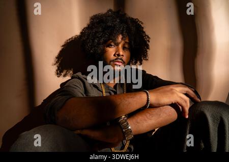 Giovane con un acconciatura afro seduto con fiducia in un ambiente studio. La sua espressione è calma e introspettiva, con luci monocromatiche vuote Foto Stock