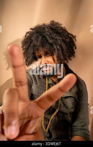 Un uomo con un'acconciatura afro posa con fiducia in un ambiente da studio, indossando una catena d'oro e mostrando un segno di pace con la mano, creando un expres Foto Stock