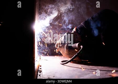 Uomo irriconoscibile impegnato nella saldatura, generando scintille brillanti e fumo. Indossando l'equipaggiamento protettivo e lo scudo, la scena cattura l'energia dinamica o Foto Stock