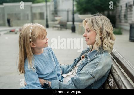 Un tenero momento di legame tra una madre e sua figlia, seduta su una panchina di legno in una tranquilla strada cittadina, condividendo un profondo legame e gioiosa Foto Stock