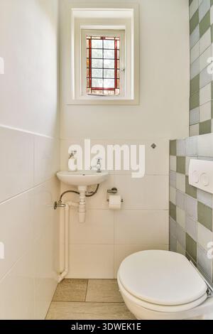 Un bagno luminoso e moderno dalle linee pulite, WC bianco e un elegante lavandino su pareti piastrellate. La finestra aggiunge luce naturale allo spazio Foto Stock