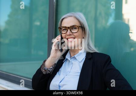 Ritratto di una donna d'affari anziana e sicura di sé che indossa gli occhiali e fa una telefonata con il suo smartphone mentre si trova all'aperto di fronte a un ufficio moderno Foto Stock