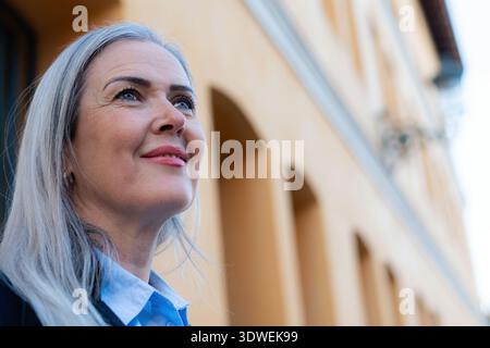 Ritratto di una donna d'affari di successo con i capelli grigi, in piedi con fiducia di fronte a un edificio moderno, sorridente e con un senso di speranza Foto Stock