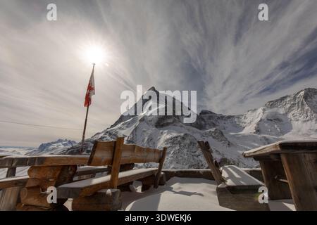 Il maestoso Cervino sotto un sole luminoso visto dalle panchine di legno del rifugio Schonbiel. Una bandiera svizzera ondeggia nel paesaggio innevato di Zermatt, Vala Foto Stock