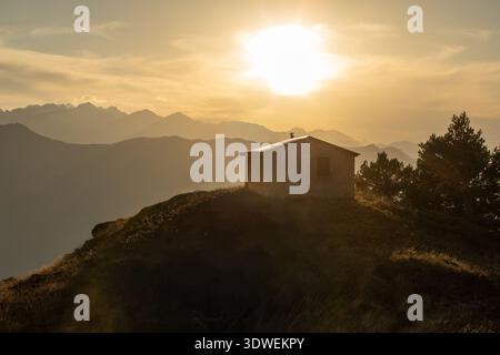 Maestoso tramonto sulle montagne dei Pirenei, che proietta un caldo bagliore su una cabina solitaria sulla cima di una collina, con sagome di pini e cime di montagna Foto Stock