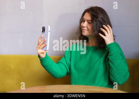 Una donna in un accogliente bar, che indossa un maglione verde, guarda il suo smartphone con un'espressione premurosa. È seduta su un divano giallo in un bancomat rilassato Foto Stock