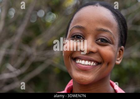 Donna afroamericana adulta sorridente mentre guarda la macchina fotografica nel parco, indossa una camicia rosa con colletto Foto Stock