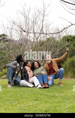 Amici diversi seduti sull'erba nel parco scattano selfie con lo smartphone, indossando sciarpe da giacca Foto Stock