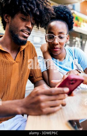 Una coppia si siede vicino a un tavolo, condividendo un momento di gioia guardando lo schermo di uno smartphone. Il loro comportamento rilassato e allegro indica un positi Foto Stock