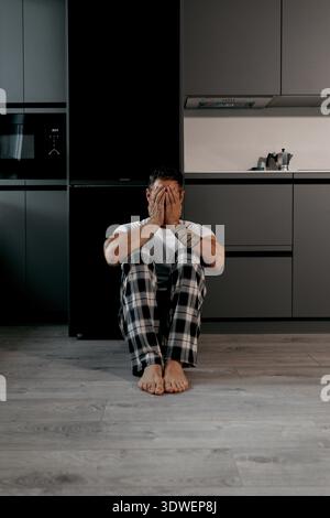 Un uomo in pigiama siede sul pavimento di una cucina con la testa in mano, trasmettendo stress o contemplazione a casa, circondato da moderni elettrodomestici e CA Foto Stock