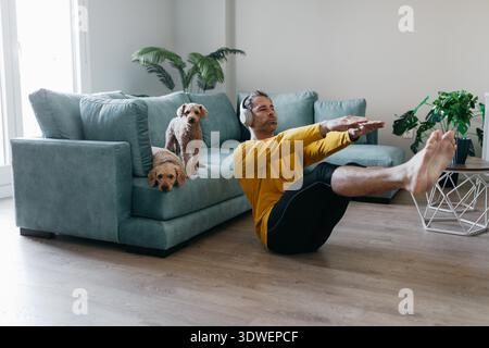 Un uomo si allena a casa, si siede sul pavimento indossando le cuffie. Due cani curiosamente lo guardano da un divano turchese, creando un c Foto Stock