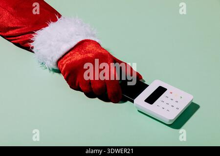 Immagine festosa di una mano in un guanto rosso di Babbo Natale con un lettore di carte bianche su uno sfondo verde chiaro, che simboleggia lo shopping natalizio e la paga moderna Foto Stock