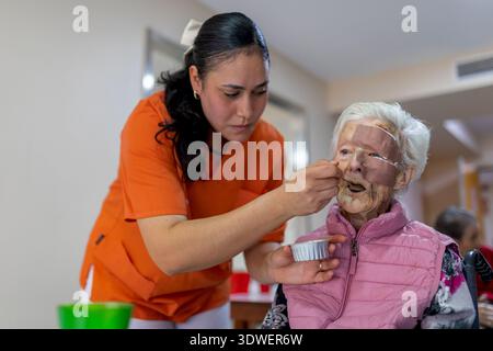 Un caregiver in uniforme arancione aiuta una donna anziana con medicazioni ferite in una residenza di anziano. Sta dando da mangiare alla donna che siede su una sedia a rotelle Foto Stock