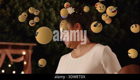 Donna matura in piedi con parte superiore bianca a manica corta rivolta verso il giardino, con sfere emoji galleggianti Foto Stock