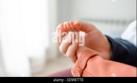 Primo piano del piede di un neonato tenuto delicatamente dalla mano di un genitore. L'immagine cattura la tenerezza e la fragilità della prima genitorialità a casa, e. Foto Stock