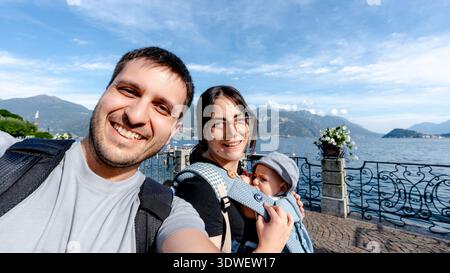 Una famiglia gioiosa si fa un selfie lungo la pittoresca passeggiata di Menaggio, sul lago di Como. Il tranquillo lago e le maestose montagne creano una ba mozzafiato Foto Stock