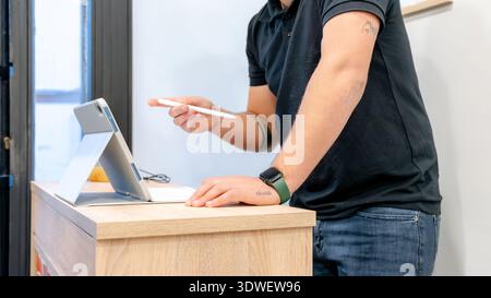 Un barbiere che indossa un abbigliamento casual interagisce con un tablet al banco della reception di un moderno barbiere. L'impostazione enfatizza l'uso della tecnologia Foto Stock