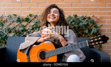 Una giovane donna con i capelli ricci si gode un momento di relax, tenendo una tazza mentre si siede con una chitarra. Trasuda calma e soddisfazione in un ambiente accogliente Foto Stock