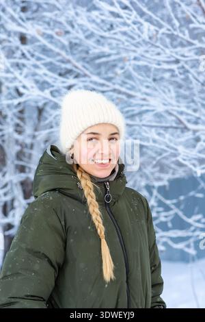 Ritratto di una donna sorridente vestita con un caldo cappotto e un cappello in maglia, in un paesaggio invernale innevato. Appare gioiosa nell'atmosfera frizzante e serena Foto Stock