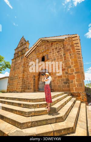 Una donna con cappello e gonna floreale fotografa la storica Cappella di Gesù Risorto, una splendida chiesa rustica nell'affascinante Barichara, Colombia, sotto Foto Stock