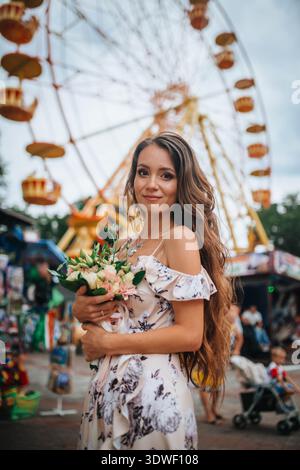 Una donna con un abito floreale regala un mazzo di fiori, davanti a una ruota panoramica colorata in una fiera vivace. La scena cattura un mix di bellezza Foto Stock