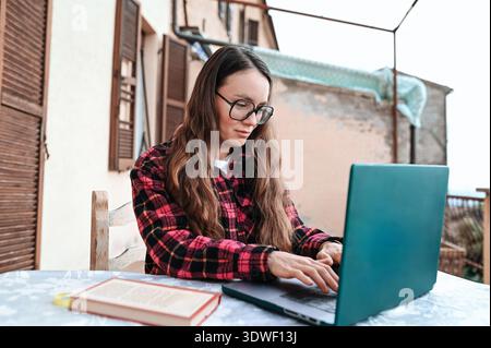 Una donna con gli occhiali e una camicia a quadri può essere indossata su un notebook mentre è seduta a un tavolo all'aperto, parte della sua routine produttiva mattutina. Un libro si trova accanto a. Foto Stock