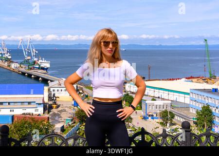 Una donna si erge con sicurezza affacciata su un porto affollato, vestita con un abbigliamento casual mentre si prende il sole. Navi da carico e gru popolano il wat Foto Stock