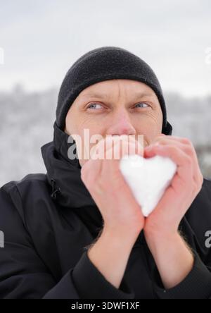Un uomo avvolto in una giacca nera e in un berretto regge una palla di neve a forma di cuore su uno sfondo innevato e sfocato. Il suo sguardo riflette il sereno inverno a. Foto Stock