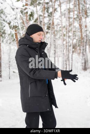 Un uomo vestito con un cappotto invernale nero e un cappello si trova in una foresta innevata, addossando i guanti. La scena cattura la bellezza serena di un paesaggio invernale Foto Stock