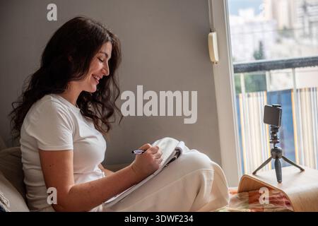 Una donna sta scrivendo un blog da casa, trasmettendo in streaming dal vivo utilizzando il suo smartphone su un cavalletto. È seduta comodamente e crea contenuti coinvolgenti per il suo pubblico Foto Stock